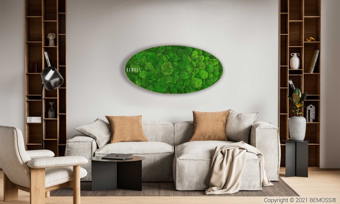 Een moderne woonkamer met een beige bank versierd met bruine en beige kussens, een witte fauteuil en twee zwarte bijzettafels. Aan de muur hangt een groen Mosschilderij Ellips BOLMOSS Lichtkunstwerk. Houten planken met decoratieve spullen flankeren de bank en links staat een staande lamp.