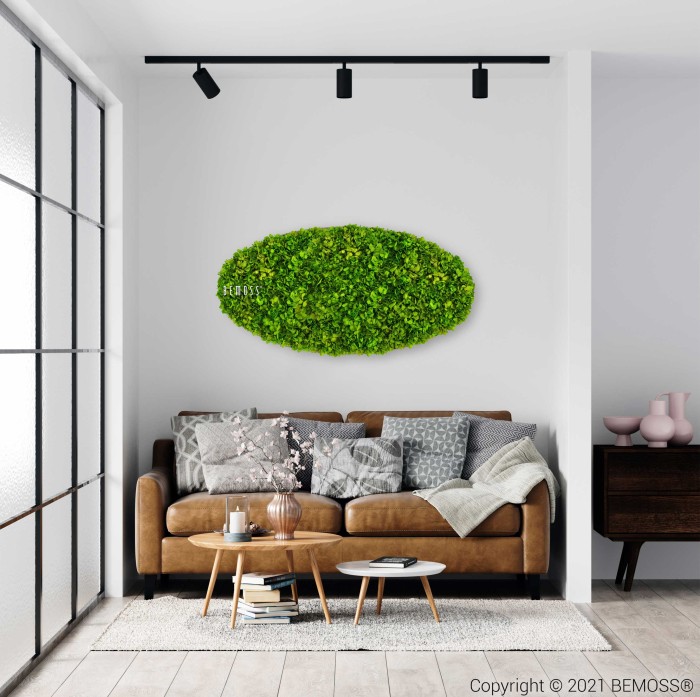 Een moderne woonkamer met een bruine bank versierd met grijze en beige kussens. Boven de bank hangt een groene Mosschilderij Ellips BEMOSS® MIMI, wat een vleugje natuur toevoegt. Op een vloerkleed staat een houten salontafel met een theepot en glazen. Een zwarte railverlichting en een dressoir met decor maken het geheel compleet.
