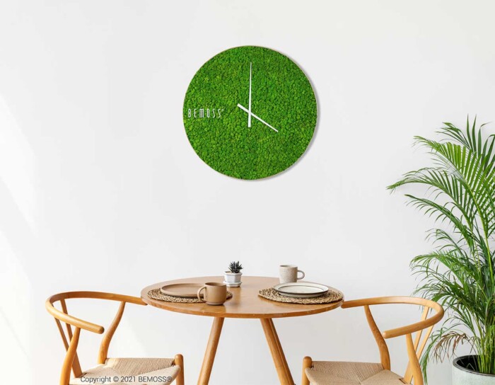 Een minimalistische eethoek is voorzien van een ronde houten tafel met twee stoelen. Op tafel staan een klein potplantje en wat serviesgoed. Op de witte muur hangt een groene Mos klok RENDIERMOS met witte wijzers en de tekst 