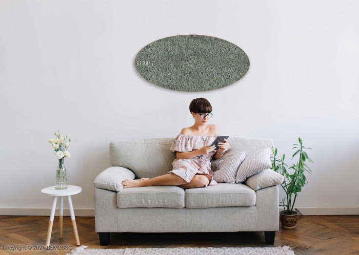 Een vrouw in een off-shoulder jurk zit op een lichtgrijze bank en leest vanaf een tablet. De kamer is minimalistisch, met een plant en een vaas met bloemen naast de bank. Aan de witte muur achter haar hangt een grote Mosschilderij Ellips RENDIERMOS mono.