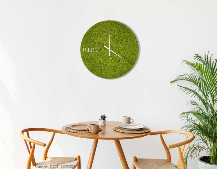 Een minimalistische eethoek met twee houten stoelen rond een ronde houten tafel. Op de tafel staat een potplant, een kom en een kopje. Boven de tafel is een groene Mos klok RENDIERMOS met witte wijzers en de merknaam 