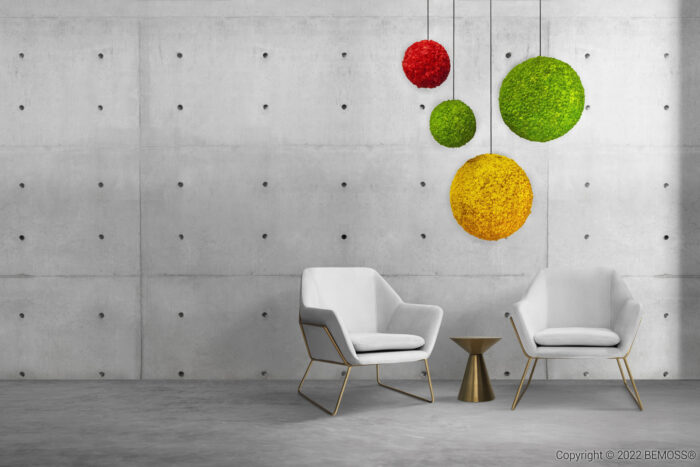 Minimalistische kamer met twee moderne witte stoelen rond een klein goudkleurig bijzettafeltje. De achtergrond bestaat uit een betonnen muur met kleine gaatjes en vier bolvormige, met mos bedekte BEMOSS-hangende decoraties in rode, gele en groene tinten.