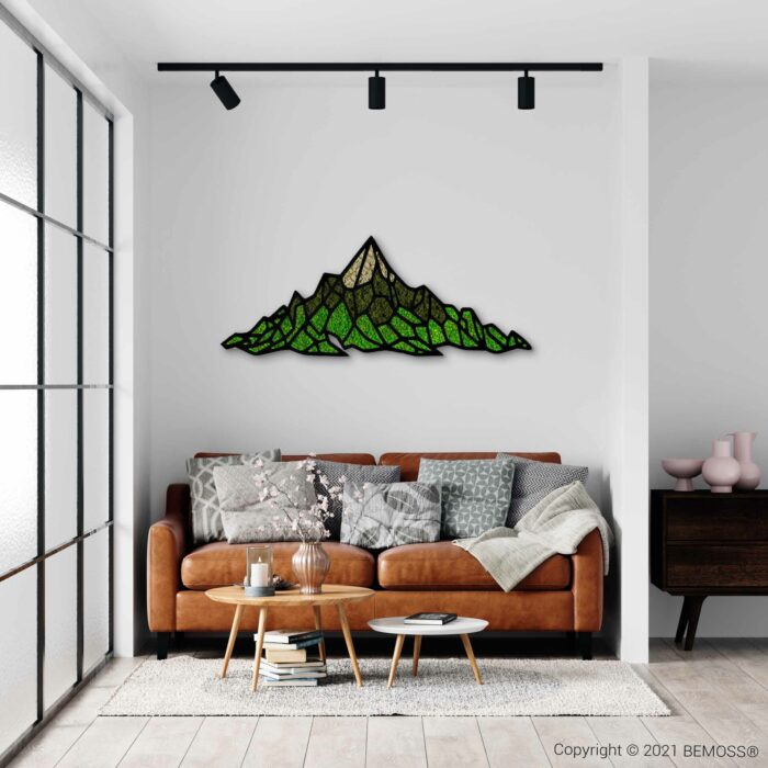 Een moderne woonkamer met een bruinleren bank versierd met verschillende kussens en een deken. Boven de bank zijn op de witte muur een glas-in-lood bergkunstwerk en een BEMOSS-mosschilderij gemonteerd. Voor de bank staat een houten salontafel met decor. Grote ramen en spoorverlichting zijn aanwezig.