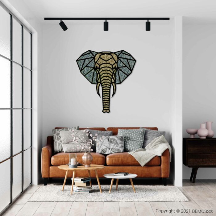 Een moderne woonkamer is voorzien van een bruinleren bank met diverse grijze kussens en kussens met patronen, en een lichte deken. Boven de bank hangt een geometrisch glas-in-lood olifantenkopkunstwerk. Een houten salontafel met decorartikelen staat op een wit vloerkleed, naast een levendige BEMOSS-mosschilderij die een weelderige groene textuur toevoegt.