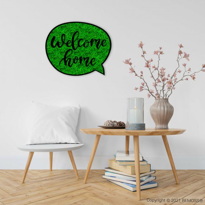 Een minimalistische woonkamer is voorzien van een witte stoel met een wit kussen op een lichte houten vloer. Op een houten bijzettafeltje staat een vaas met roze bloemen, een glazen kandelaar en een stapel boeken. Op een opvallende BEMOSS-mosschilderij aan de muur staat in decoratief schrift 