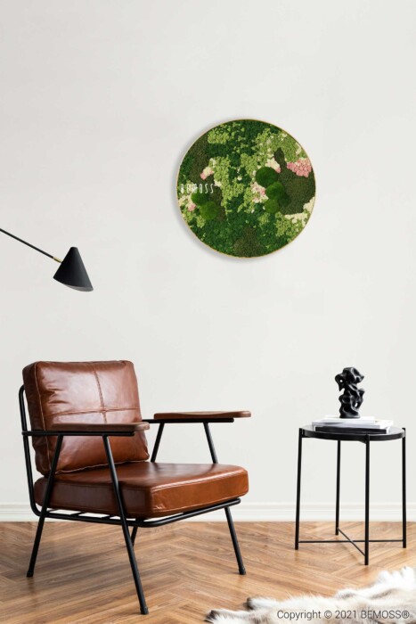 Een moderne woonruimte is voorzien van een bruinleren stoel met een zwart frame, een klein zwart bijzettafeltje met een sculptuur en een zwarte, schuine vloerlamp. Aan de witte muur boven de lichtgekleurde houten vloer hangt een Mosschilderij cirkel BEMOSS® ORTHO PINK, wat een organische toets aan de kamer geeft.