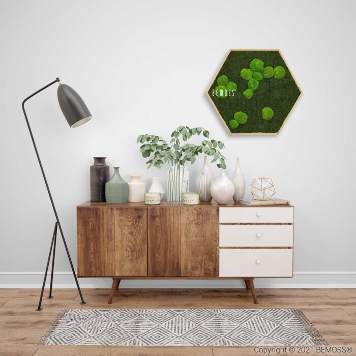Een modern interieurtafereel met een houten consoletafel uit het midden van de eeuw tegen een witte muur. Op de tafel staan diverse decoratieartikelen, waaronder vazen, kaarsen en een plant. Boven, aan de muur, staat een achthoekige BEMOSS-mosschilderij. Links staat een vloerlamp op een tapijt met patroon.