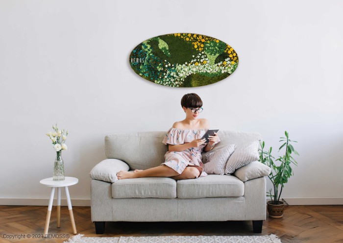Een vrouw in een off-shoulder jurk zit op een beige bank een boek te lezen. De minimalistische kamer is voorzien van een klein rond wit bijzettafeltje met een plant, een potplant op de vloer en een ovaalvormige groene Mosschilderij Ellips FLATMOSS boven de bank.