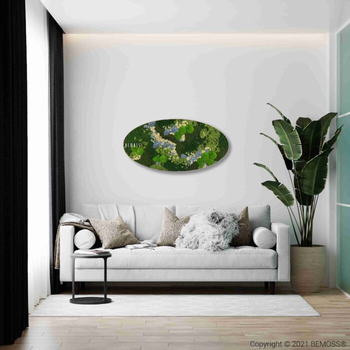 Een moderne woonkamer met een witte bank, verschillende kussens, een klein bijzettafeltje, een grote potplant en de Mosschilderij cirkel BEMOSS® ORTHO ICE - een ovaal groen kunstwerk - aan de muur boven de bank.