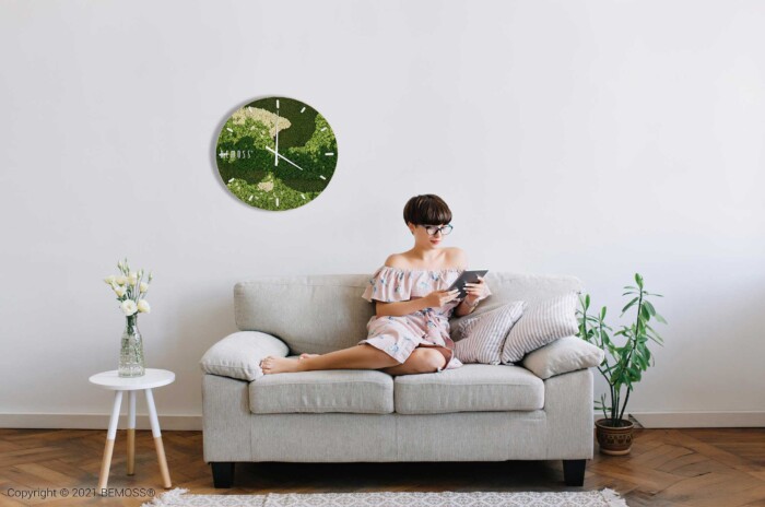 Een persoon die een lichtgekleurde off-shoulder jurk draagt, zit op een beige bank een tablet te lezen. Links staat een tafeltje met een vaas met bloemen, en de muur erboven siert een grote groene klok. De moderne setting is voorzien van weelderige planten op de vloer en een elegante Mos klok BEMOSS® ORTHO SPRING met wijzerplaat die de ruimte siert.