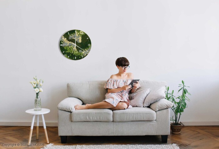 Een vrouw zit op een beige bank in een minimalistische woonkamer een boek te lezen. Ze draagt een bril en een off-shoulder jurk. Aan de muur boven haar hangt een ronde Mos klok BEMOSS® ORTHO SPRING met wijzerplaat. Naast de bank staat een tafeltje met een vaas en een kamerplant.