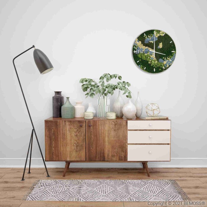 Een houten dressoir met witte laden staat tegen een witte muur, bedekt met vazen, een plant en decor. Links staat een zwarte staande lamp. Daarboven hangt de Mosschilderij cirkel BEMOSS® ORTHO ICE. Een vloerkleed met patroon bedekt de vloer.