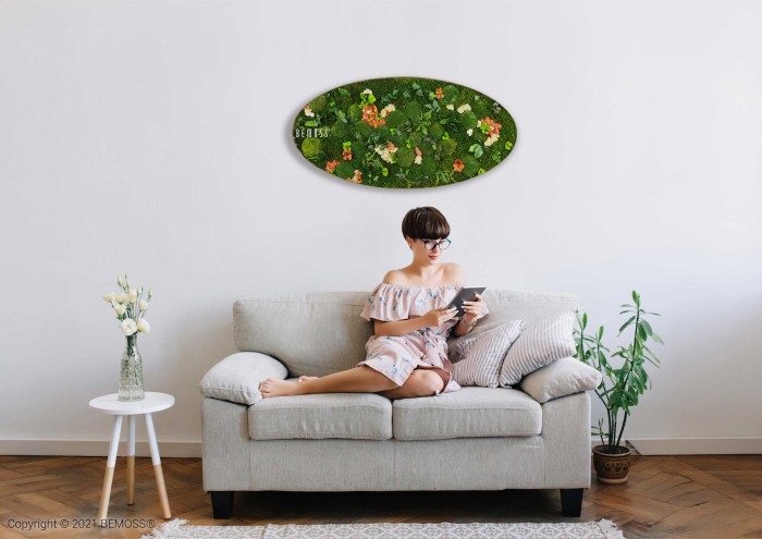 Een vrouw in een off-shoulder bloemenjurk zit op een lichtgrijze bank een boek te lezen. De bank staat in een minimalistische kamer met witte muren, een groene plant op de vloer en een ronde tafel met een vaas met bloemen. Boven de bank hangt een ovale Mosschilderij Ellips FLATMOSS.