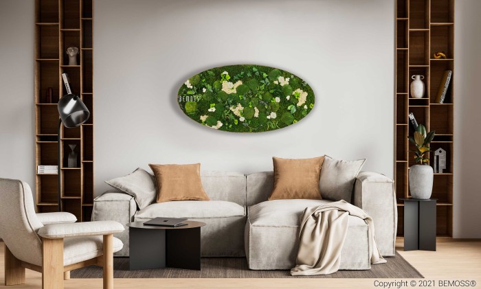 Een moderne woonkamer is voorzien van een lichtgrijze bank versierd met beige en grijze kussens en een gezellige deken. Boven de bank hangt een ovaal Mosschilderij Ellips FLATMOSS groen bloemenkunstwerk aan de muur. De kamer is voorzien van houten boekenplanken aan weerszijden, een zwart bijzettafeltje, een zwarte vloerlamp en een beige fauteuil.