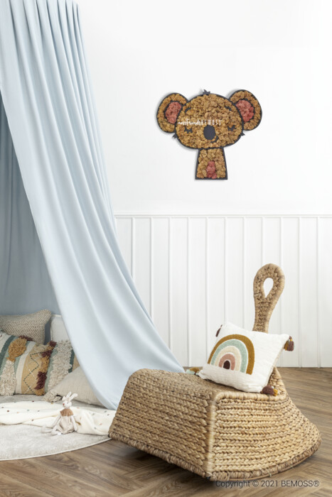 Een gezellige kinderkamer is voorzien van een rieten fauteuil met een regenboogkussen en een gehaakte pop. Ernaast hangt een lichtblauwe baldakijn, en daarboven voegt een aan de muur gemonteerde koalabeervormige decoratie een speels tintje toe. Het betoverende decor wordt compleet gemaakt door een levendige BEMOSS-mosschilderij aan de muur.