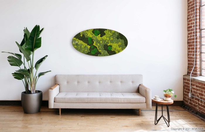 Een minimalistische woonkamer met een lichtgrijze bank tegen een witte muur, links een groene potplant en rechts een ronde tafel met een plant en boeken. Boven de bank hangt een ovaalvormige Mosschilderij Ellips BEMOSS® ORTHO TORRES. Natuurlijk licht stroomt naar binnen via een raam aan de rechterkant.