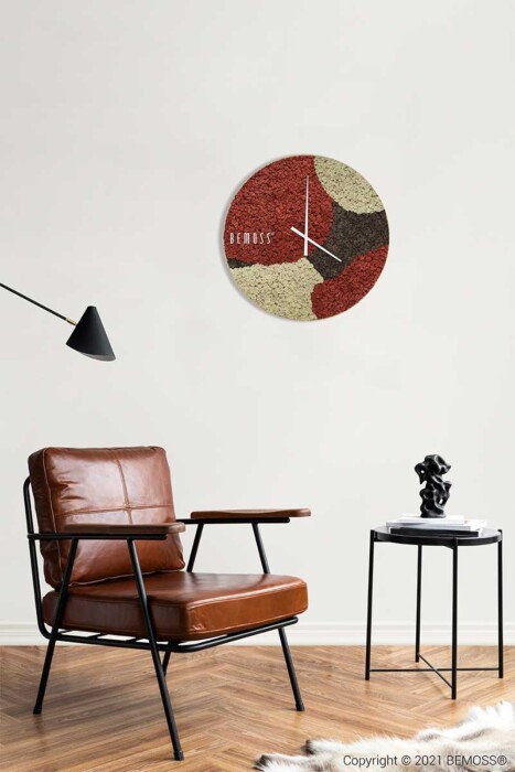 Een minimalistische woonkamer is voorzien van een bruinleren stoel met een zwart metalen frame, een klein zwart bijzettafeltje met een moderne sculptuur en een aan de muur gemonteerde structuurklok met abstracte patronen in rood, groen en beige. Een Mos klok BEMOSS® ORTHO SIENNA geeft een organische toets aan het decor, terwijl een zwarte schuine lamp aan het plafond hangt.