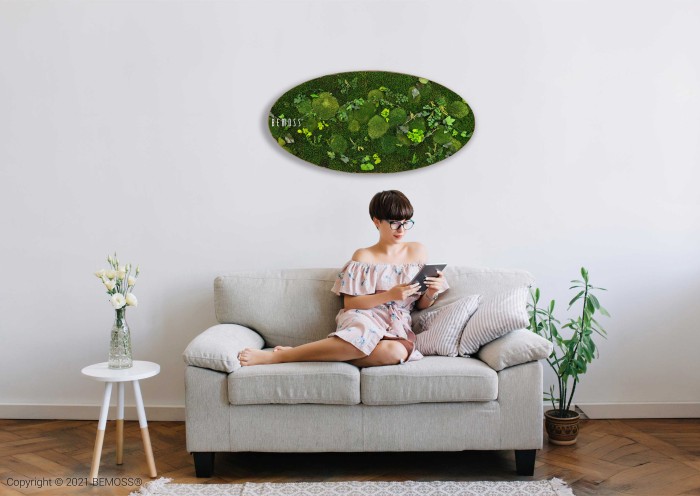 Een vrouw in een gebloemde jurk zit op een lichtgrijze bank en houdt een tablet vast. Boven haar hangt een ovaalvormige Mosschilderij Ellips PLANTEN Mesia. Links een klein bijzettafeltje met een vaas met bloemen, en rechts een potplant op de grond. De kamer heeft een minimalistische esthetiek.