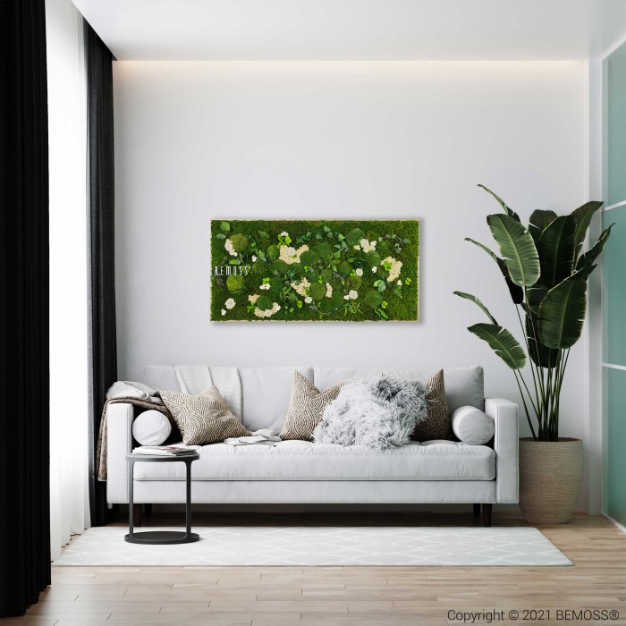Een moderne woonkamer is voorzien van een lichtgrijze bank versierd met beige en grijze kussens en een donzige grijze plaid. Boven de bank hangt een Mosschilderij PLANTEN Mesia Natural kunstwerk aan de muur. Rechts staat een grote potplant en zwarte gordijnen omlijsten een groot raam.