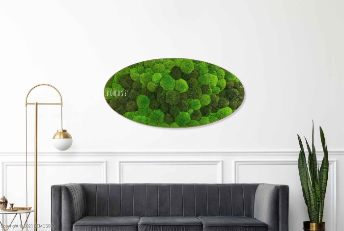 Een moderne woonkamer met een grijze bank met daarnaast een hoge groene plant. Boven de bank hangt een ovaalvormige Moshexagon PLANTEN Lomo in verschillende tinten groen. Links staat een witte en gouden vloerlamp. De muur is wit met sierlijke lijstwerk.