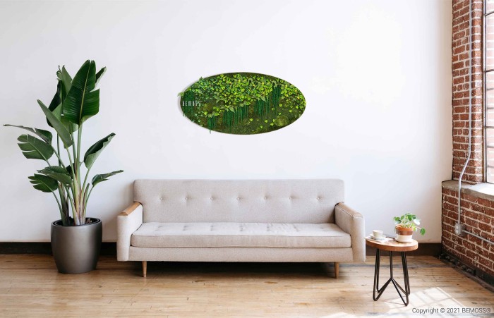 Een moderne woonkamer met een beige bank tegen een witte muur. Boven de bank hangt een ovaalvormig kunstwerk dat een weelderig groen landschap voorstelt. Links van de bank staat een grote potplant en rechts een klein rond houten tafeltje met een plant en een mok erop. Het middelpunt is echter ongetwijfeld de Moshexagon PLANTEN Lomo die charme aan de ruimte toevoegt.