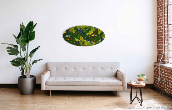 Een moderne woonkamer is voorzien van een beige bank tegen een witte muur. Links staat een grote potplant, terwijl erboven een ovaalvormig kunstwerk aan de muur hangt met een kleurrijk, abstract bloemdessin. Rechts van de bank staat een klein houten bijzettafeltje met een plant naast een Mosschilderij PLANTEN Lomo.