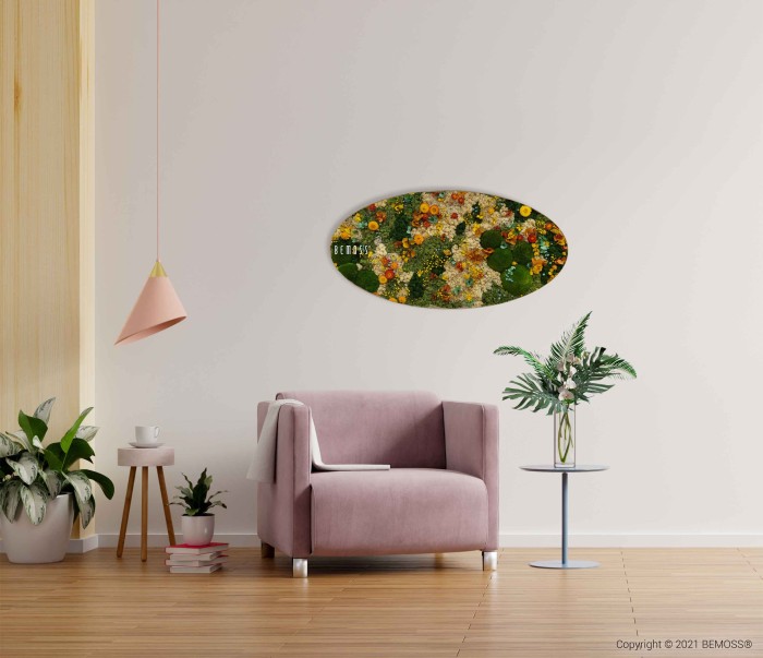 Een moderne woonkamer is voorzien van een lichtroze fauteuil, een ronde bijzettafel met een potplant en een Mosschilderij Ellips BOLMOSS DUO Natural Green aan de muur met een kleurrijk bloementafereel. Er staat nog een potplant op de vloer en een hangende hanglamp met een roze kap.