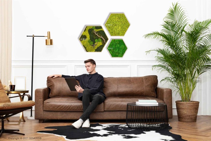 Een man zit op een bruinleren bank een tablet te lezen in een moderne woonkamer. De ruimte is voorzien van een hoge groene plant, zeshoekige kunst aan de muur, Moshexagon BEMOSS® ORTHO BORJA, een staande lamp, een koeienhuidkleed en een klein bijzettafeltje met een wit en gouden kopje.