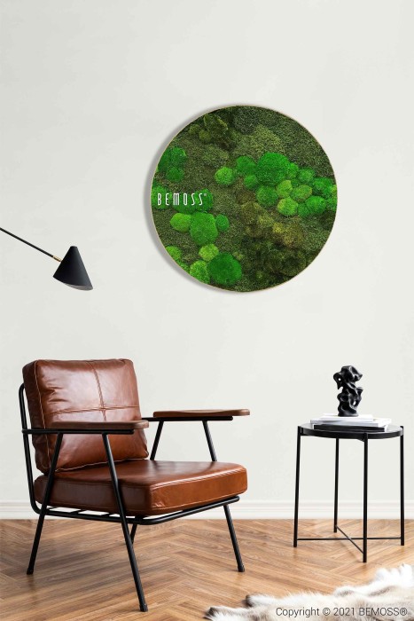 Een moderne woonkamer is voorzien van een bruinleren stoel, een zwarte bijzettafel met een beeldhouwwerk en een minimalistische zwarte wandlamp. Aan de witte muur hangt een ronde Mosschilderij Ellips BOLMOSS DUO Natural Green. De kamer heeft een houten vloer en een zacht wit vloerkleed. (© 2021 BEMOSS®).