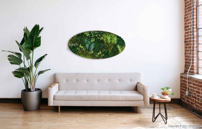 Een minimalistische woonkamer is voorzien van een lichtgrijze bank, een kleine houten salontafel met planten en een grote potplant aan de linkerkant. Boven de bank hangt een ovaalvormige BEMOSS moschilderij. Bakstenen muren en een groot raam zorgen voor een luchtige sfeer.