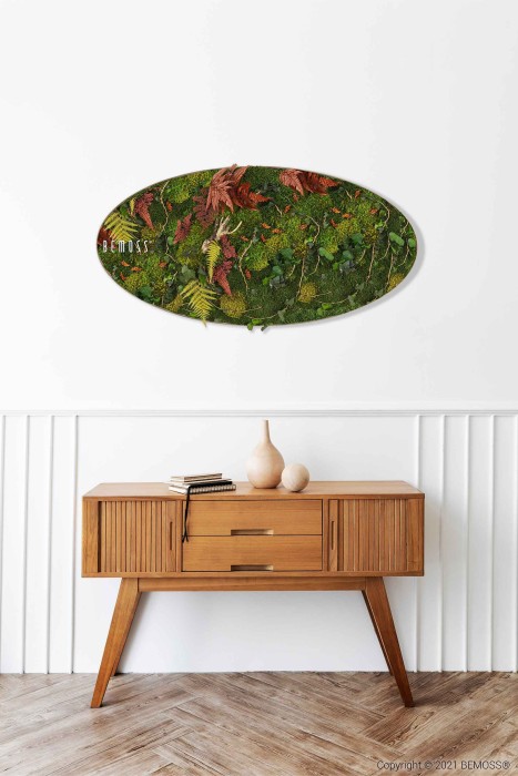 Een moderne houten consoletafel staat tegen een lichtgekleurde muur met witte lambrisering. Aan de muur boven de tafel hangt een prachtige BEMOSS-mosschilderij met groen mos, varens en rode bladeren. Op de tafel staan boeken en decoratieve vazen.