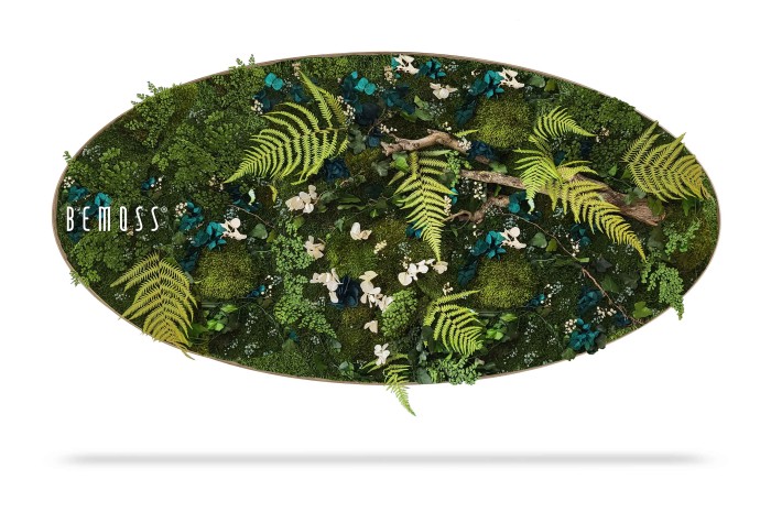 Een ovale groene muurkunst, een prachtige BEMOSS moswand, met een assortiment bewaarde planten, varens, mossen en bloemaccenten. De weelderige compositie heeft verschillende tinten groen met vleugjes witte en blauwe bloemen, waardoor een levendige, natuurlijke esthetiek ontstaat. Aan de linkerkant verschijnt het woord 