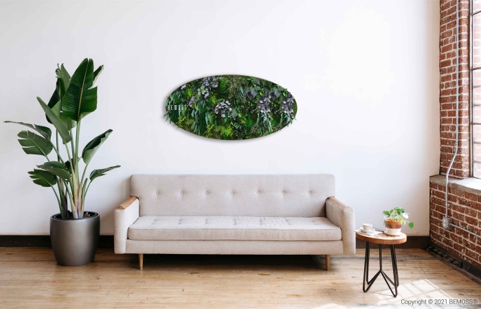 Een minimalistische woonkamer met een lichtgrijze bank tegen een witte muur. Boven de bank bevindt zich een groene, ovaalvormige BEMOSS-mosschilderij met levendige planten. Links staat een hoge potplant en rechts een houten bijzettafeltje met een plantje en een mok. Een groot raam met zichtbare stenen laat natuurlijk licht binnen.