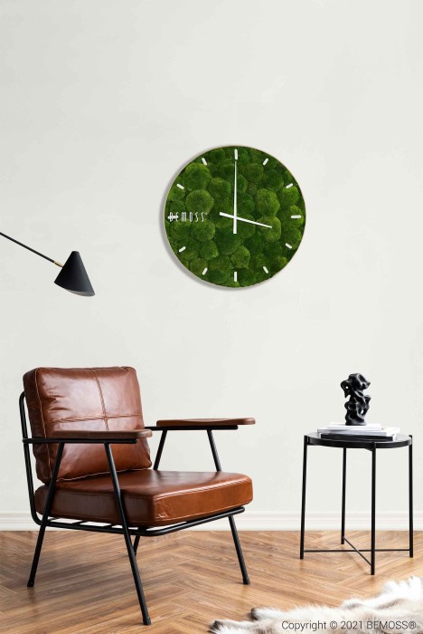 Een moderne woonruimte met een bruinleren fauteuil op een houten vloer met een wit, harig vloerkleed. Aan de wand hangt een Mos klok BOLMOSS Minerva met wijzerplaat. Naast de stoel staat een zwart bijzettafeltje met een klein beeldje en boeken, terwijl links een zwarte wandlamp is gemonteerd.