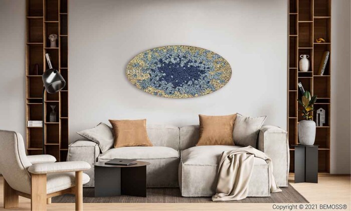 Een moderne woonkamer is voorzien van een lichtgekleurde bank met beige en bruine kussens. De achterwand heeft een groot, ovaal abstract kunstwerk. Aan elke kant van de bank staan houten planken met diverse decoratieve items. Links staat een gezellige fauteuil, aangevuld met een levendige BEMOSS-mosschilderij aan de muur.