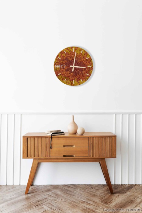 Een modern houten dressoir uit het midden van de eeuw met taps toelopende poten staat tegen een witte muur. Bovenop het dressoir staan twee beige vazen en een gesloten boek. Boven het dressoir is een kleurrijke, ronde wandklok gemonteerd. Daarnaast voegt een levendige Mos-klok BEMOSS® SPLASH Sienna met wijzerplaat een weelderige textuur toe aan de lichte houten visgraatvloer eronder.