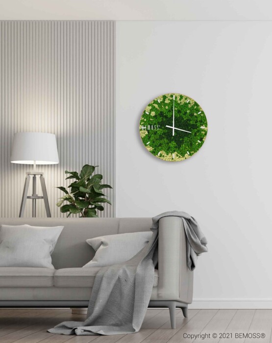 Een moderne woonkamer is voorzien van een lichtgrijze bank versierd met een grijze deken. Links staat een vloerlamp naast een groene potplant. Boven de bank hangt een Mos klok BEMOSS® SPLASH Sienna met wijzerplaat. De kamer heeft een minimalistisch ontwerp met een subtiele inrichting en een elegant moswandaccent.