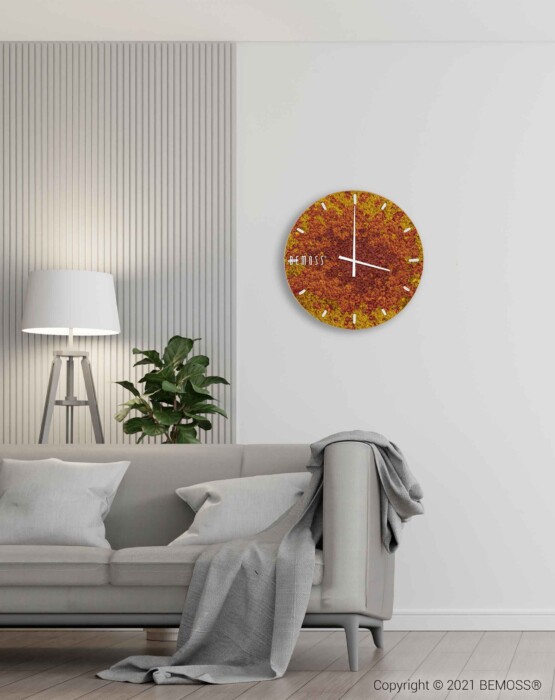 Een moderne woonkamer is voorzien van een grijze bank met een deken over de armleuning, een potplant en een staande lamp. Een oranje wandklok met structuurontwerp hangt aan een witte muur met verticale paneeldetails. In de buurt voegt een Mos klok BEMOSS® SPLASH Sienna met wijzerplaat een natuurelement toe aan het decor.