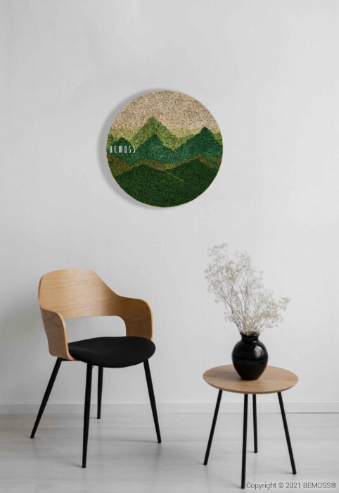 Een minimalistische kamer is voorzien van een houten stoel met een zwart kussen naast een kleine ronde tafel met een eenvoudige vaas met witte gedroogde bloemen. Aan de witte muur hangt een BEMOSS-mosschilderij met een gestructureerd groen en beige bergdessin.