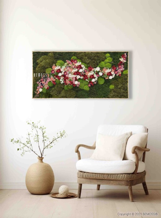 Een gezellig hoekje met een houten fauteuil met witte kussens op een lichte houten vloer. Ernaast staat een grote beige vaas met daarin een plant met kleine blaadjes. Aan de muur erboven hangt een levendige Mosschilderij PLANTEN Acebo BLUE met groen mos en trossen roze en witte bloemen.