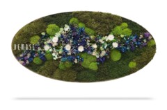 Een ovaalvormig bloemstuk bevat een verscheidenheid aan bloemen en planten. Het middelpunt is een mix van groen mos en witte, blauwe en paarse bloemen. Het woord "BEMOSS" wordt aan de linkerkant van het arrangement weergegeven.