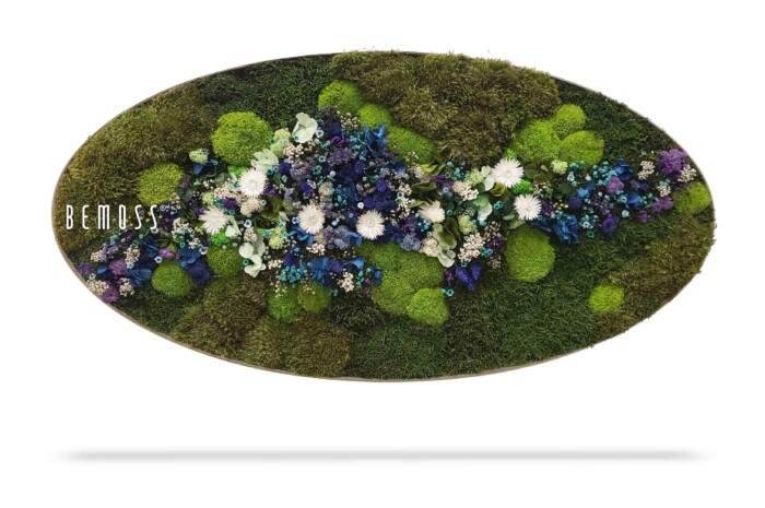 Een ovaalvormig bloemstuk bevat een verscheidenheid aan bloemen en planten. Het middelpunt is een mix van groen mos en witte, blauwe en paarse bloemen. Het woord 