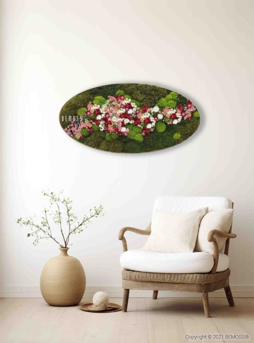 Een zitkussen en kussen liggen op een houten fauteuil in een kamer met beige muren. Boven de stoel hangt een groot, ovaalvormig stuk muurkunst met mos en kleurrijke bloemen. Een hoge vaas met delicate takken staat op de vloer naast de stoel.