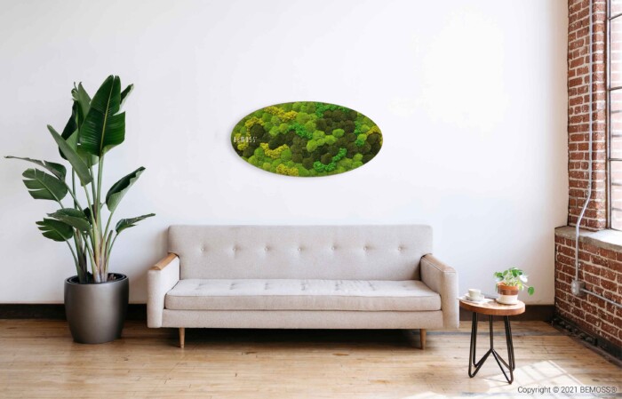 Een moderne woonkamer met een lichtgrijze bank, een salontafel met houten poten en planten erop, een grote potplant links en een ovaalvormig groen moskunstwerk op de witte muur boven de bank. Een hoog, smal raam is rechts.