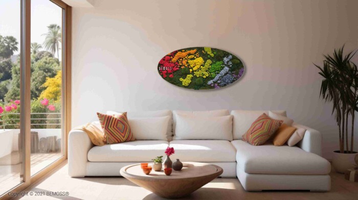 Een lichte woonkamer heeft een grote witte bank, kleurrijke kussens, een ronde houten salontafel met kleine plantjes, een groot raam en een ovaal Mosschilderij cirkel BEMOSS® ORTHO IRIS wandkunstwerk met een levendig mosdesign.