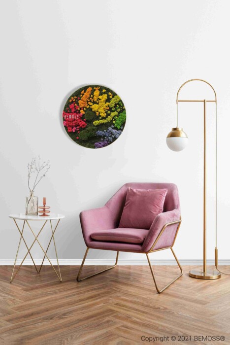 Een moderne woonkamer is voorzien van een roze fluwelen fauteuil, een ronde bijzettafel met decor, een hoge staande lamp en de Mosschilderij cirkel BOLMOSS DUO Natural Green muurkunst met kleurrijk bloemen- en groen mosdesign op een witte muur.