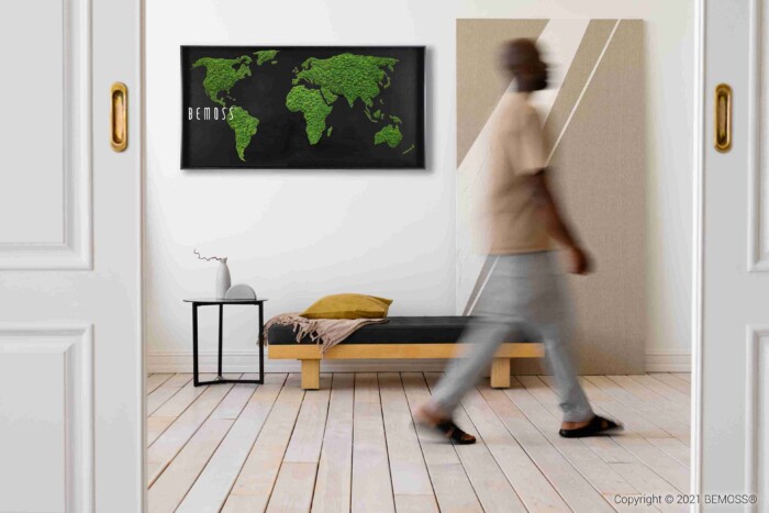 Een wazig persoon loopt langs een minimalistische slaapkamer met een laag houten bed, een klein bijzettafeltje, een kussen en de Mosschilderij Wereldkaart RONDA zwart - een kunstwerk van een wereldkaart met groene landmassa's op een zwarte achtergrond - dat aan de muur hangt.